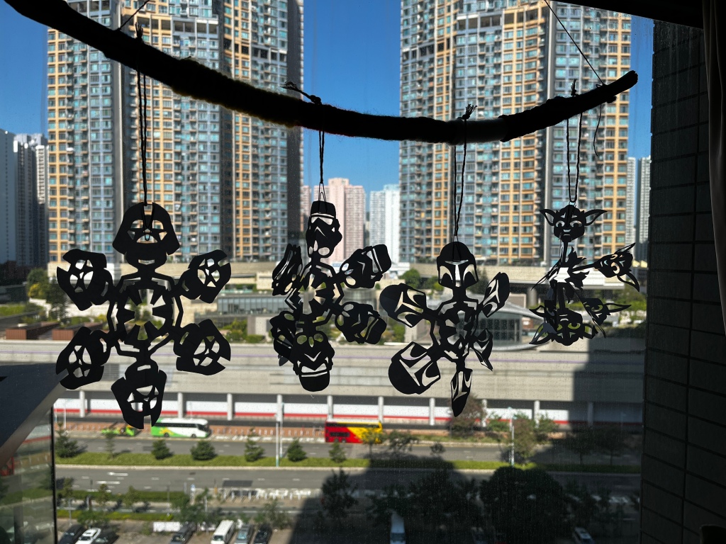 String of Star Wars snowflakes hang up from a beam of yarn covered wood against a window. Skyline of Hong Kong in the background. Idea is for Christmas decoration inspiration. 